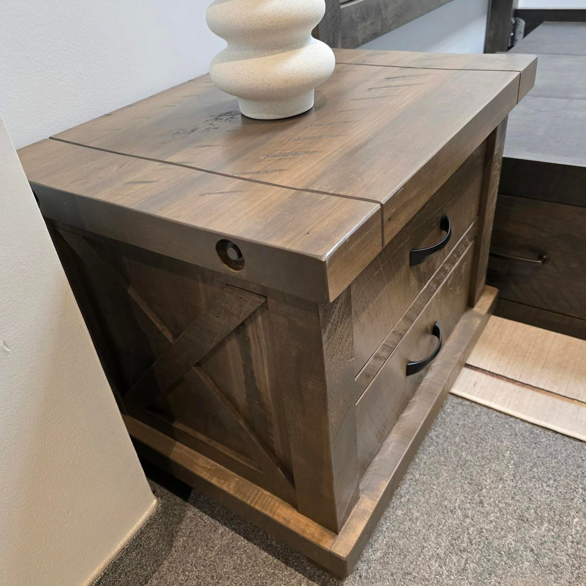 A rustic dark brown wooden nightstand with two drawers and black metal handles, shown in a bedroom setting next to a bed frame and a white decorative vase.