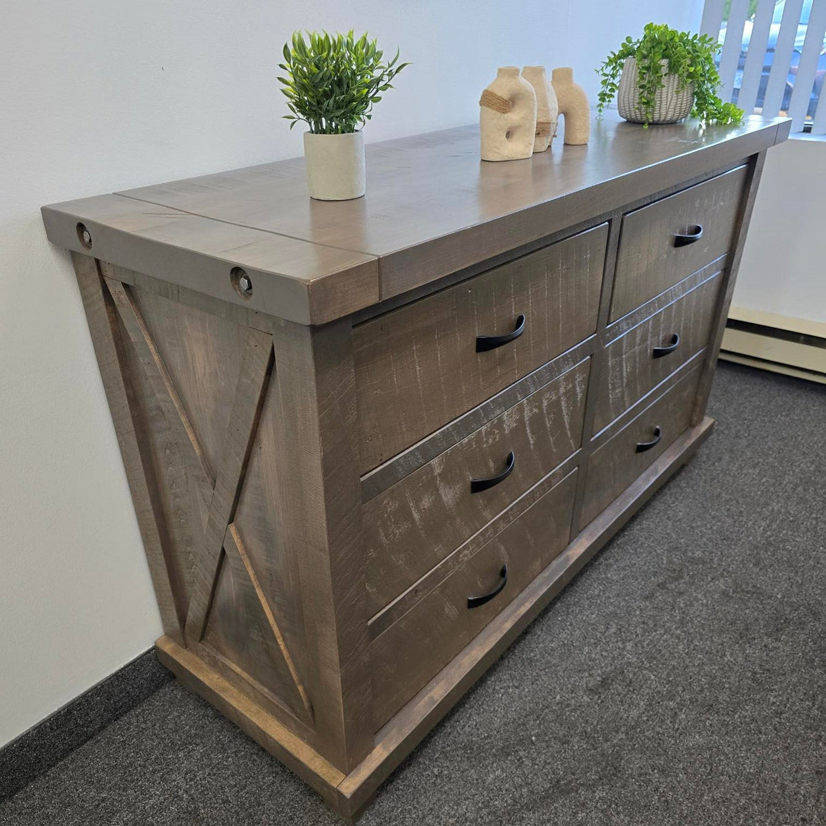 A rustic gray-brown wooden dresser with six drawers and black metal handles, styled with small potted green plants and beige ceramic vases on top, set against a white wall and gray carpet.