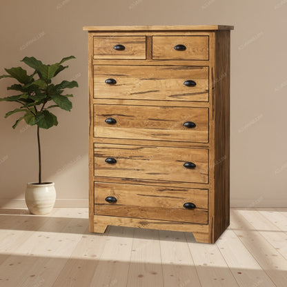 A rustic wooden 6-drawer chest of drawers with black metal knobs, set against a beige wall and light wood floor next to a potted green plant.
